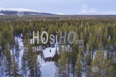 Aerial Drone Photo Of Wooden Cabin In The Remote Forest, With Snow Covered Trees Landscape And Mountains In Lapland, Finland